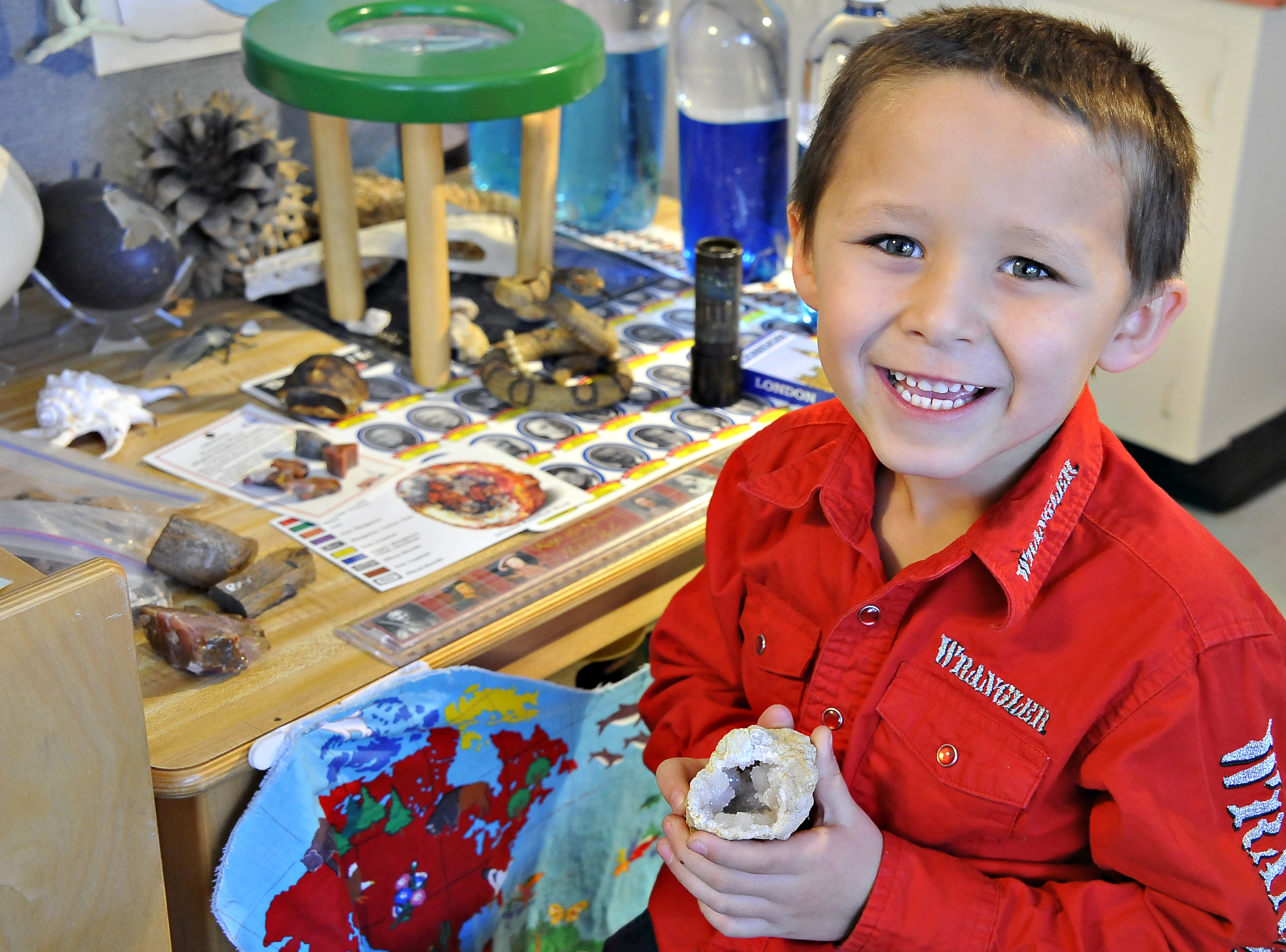 A little boy in a red shirt smiles and holds up a rock