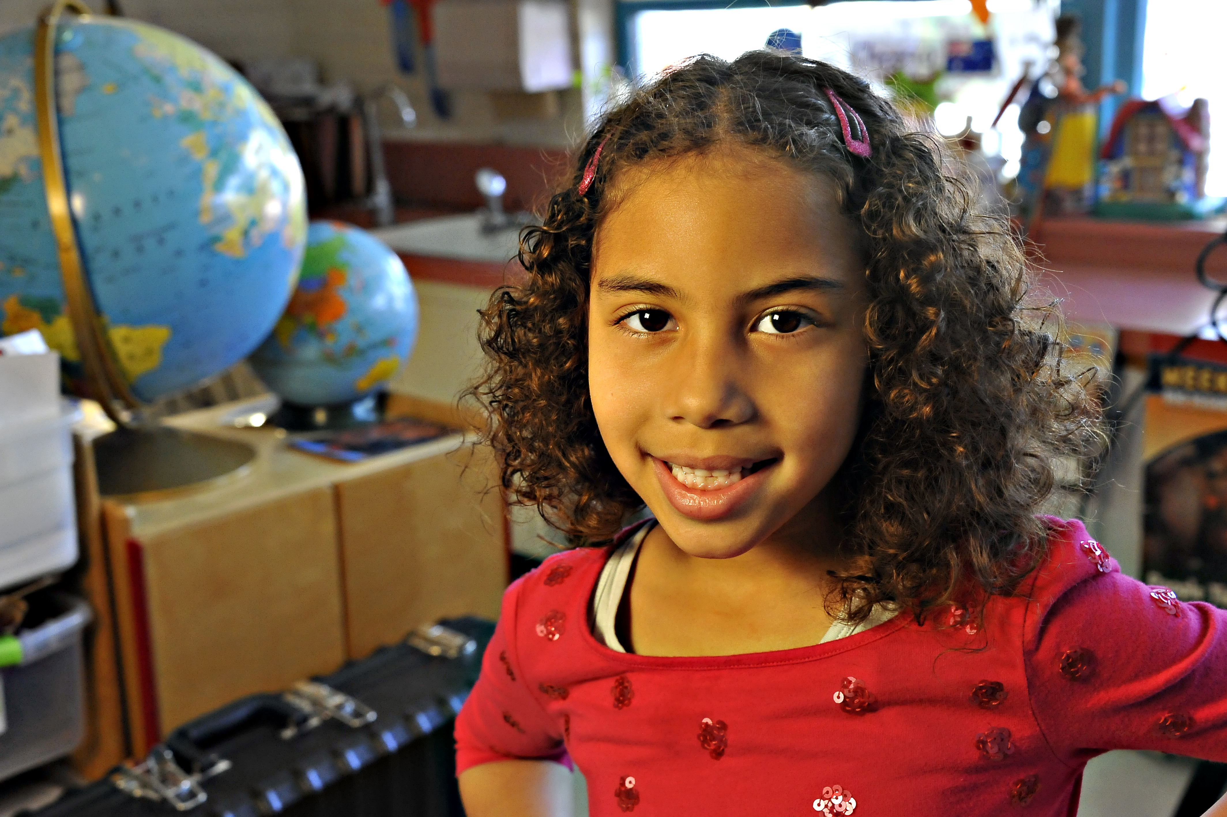 A little girl in a red shirt smiles in front of two globes in the classroom